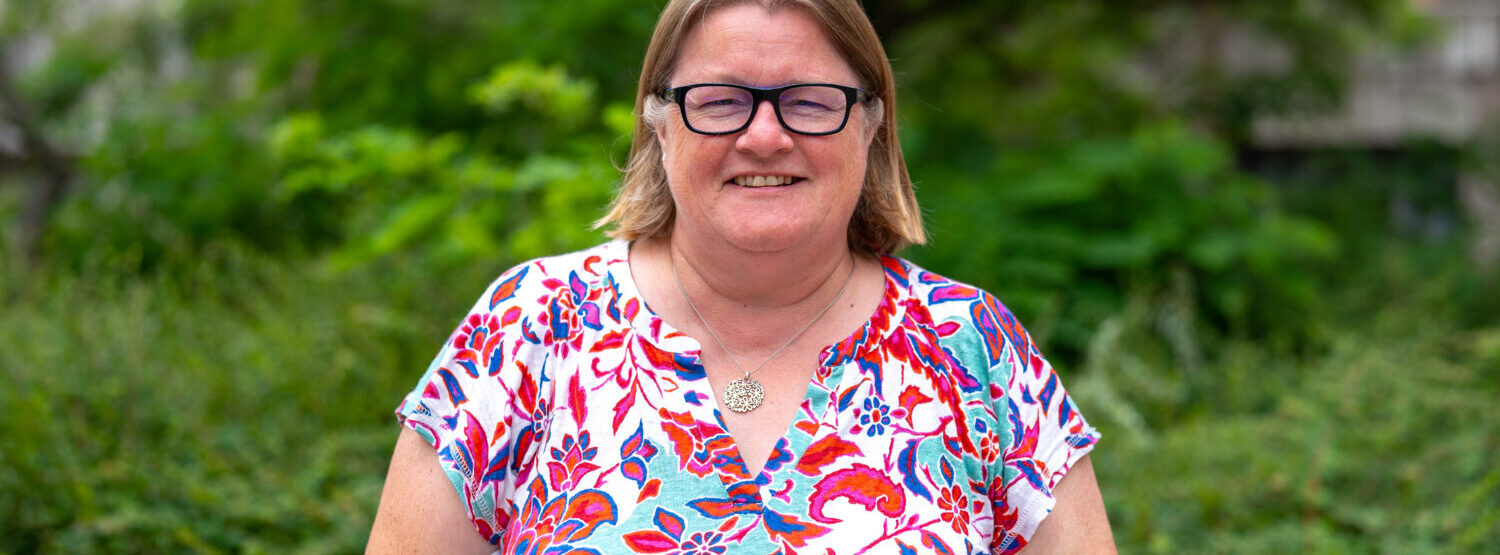A woman wearing glasses and a colourful top, smiling. Outside.