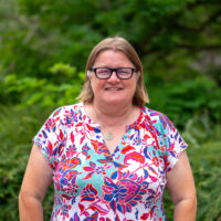 A woman wearing glasses and a colourful top, smiling. Outside.
