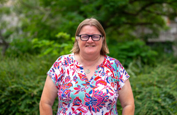 A woman wearing glasses and a colourful top, smiling. Outside.