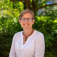 A woman with short hair and glasses, smiling. Outside.