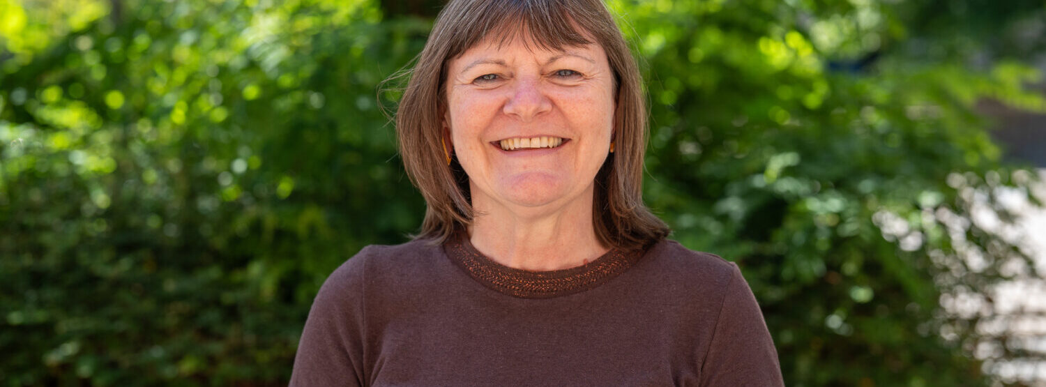 A woman with short brown hair wearing a brown top, smiling. Outside.
