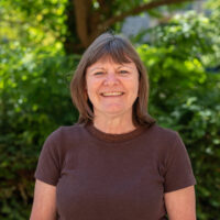 A woman with short brown hair wearing a brown top, smiling. Outside.