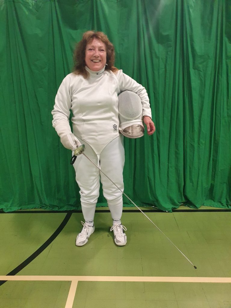 Susan in her fencing uniform, smiling in front of a green curtain. 