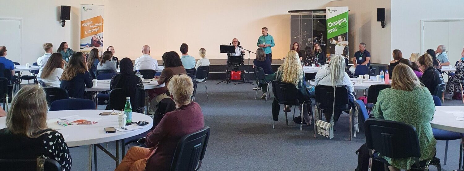 A speaker stood at the front of a room, with a presentation behind them. People are sat at round tables watching.