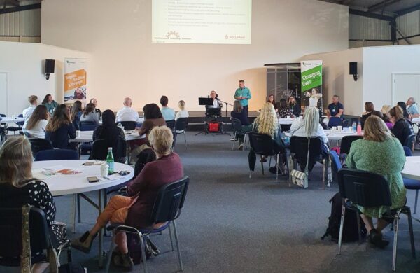 A speaker stood at the front of a room, with a presentation behind them. People are sat at round tables watching.