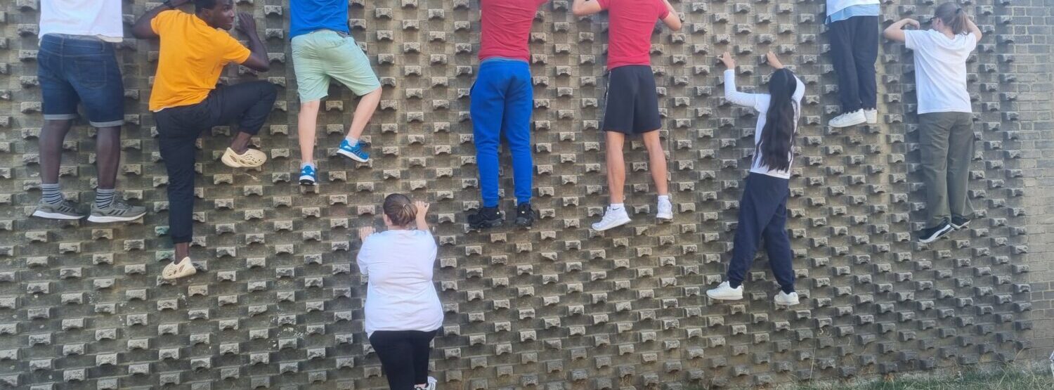 A group of young people in brightly coloured t-shirts. They are climbing up a wall, at various stages.