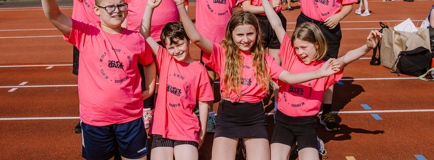 A group of children and young people in bright pink T-shirts. They are waving their arms and cheering. Taken at the Hampshire School Games 2023.