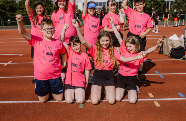 A group of children and young people in bright pink T-shirts. They are waving their arms and cheering. Taken at the Hampshire School Games 2023.