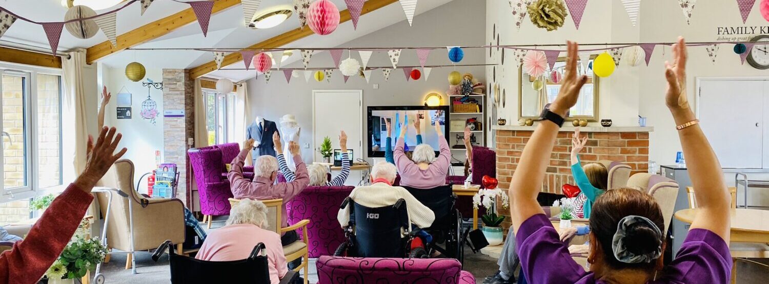 A group of older people taking part in chair based physical activity. They're in a colourful setting with bunting across the room.