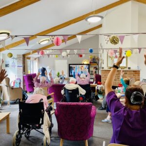 A group of older people taking part in chair based physical activity. They're in a colourful setting with bunting across the room.