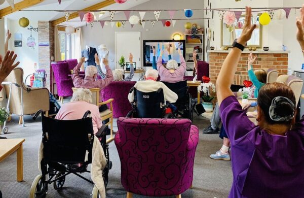 A group of older people taking part in chair based exercises in a care home. They have their arms raised above their heads. The room is very colourful with bunting hanging from the ceiling.