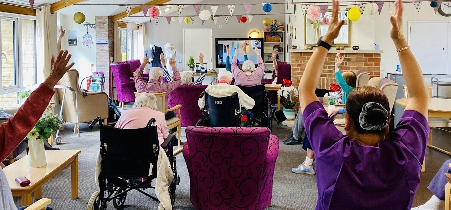 A group of older people taking part in a chair based exercise class in a care home.