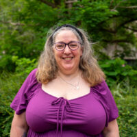 A woman wearing glasses and a purple top, smiling. Outside.