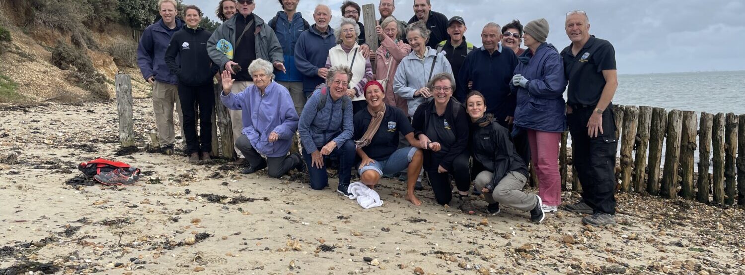 A group of 25 people from Dementia Friendly Alton, smiling at he camera at Lepe Beach, New Forest.