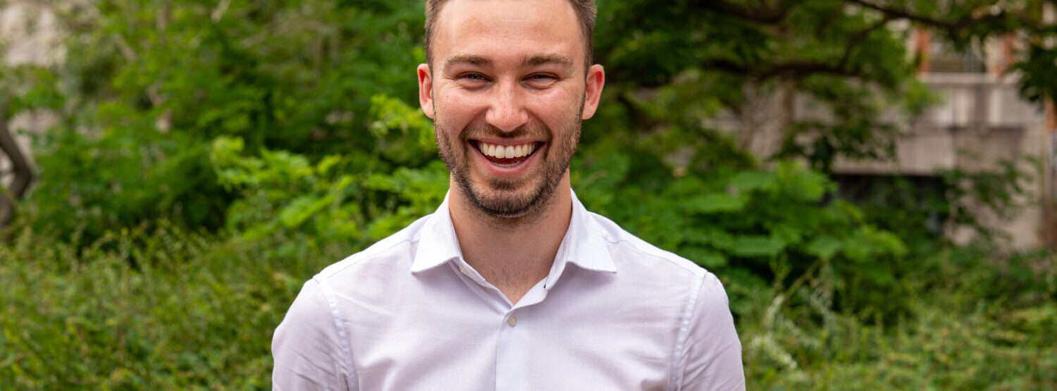 A man with brown hair and a beard in a white shirt, smiling. Outside.