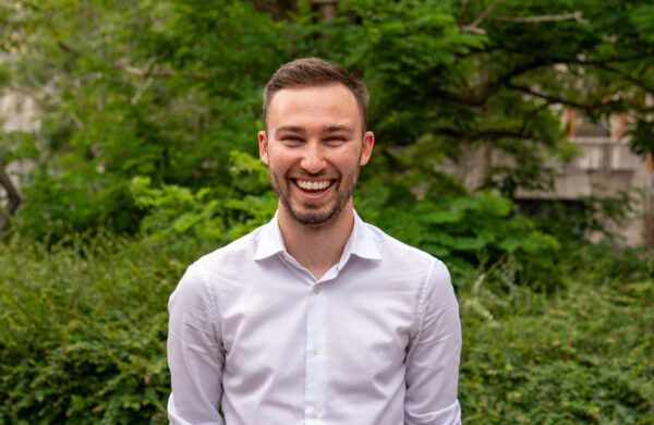 A man with brown hair and a beard in a white shirt, smiling. Outside.
