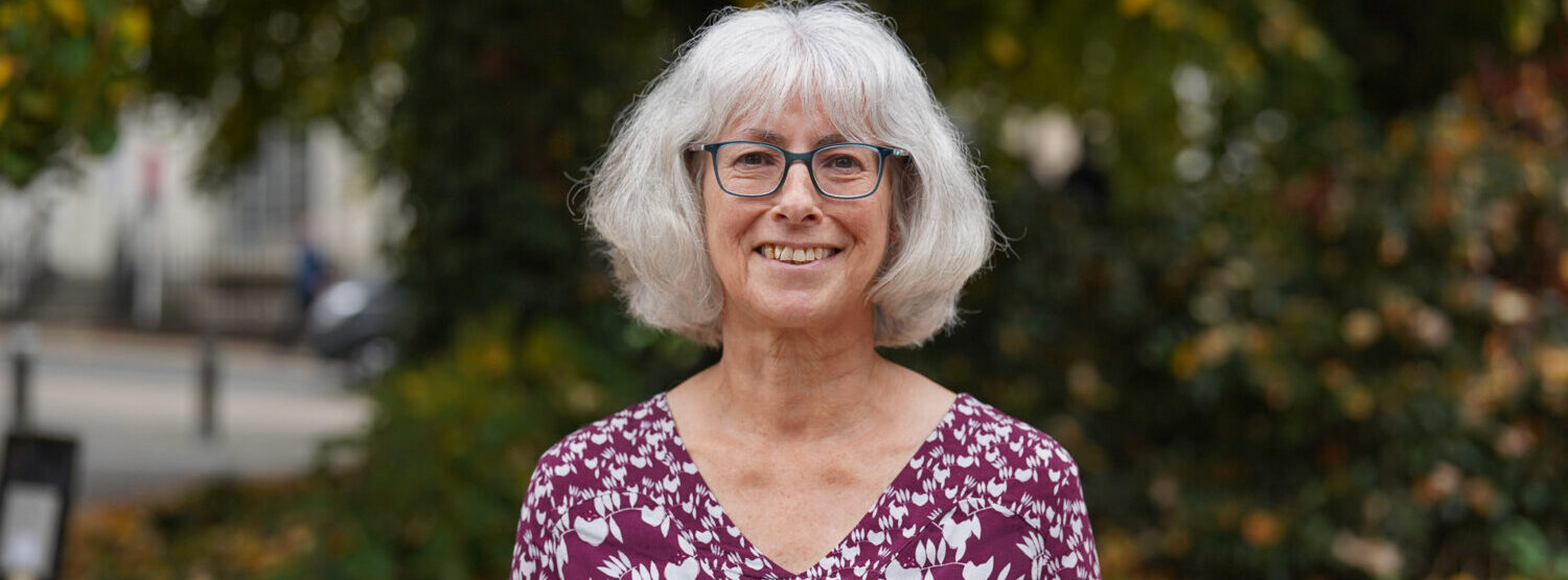 A woman in a purple blouse, wearing glasses, outside.