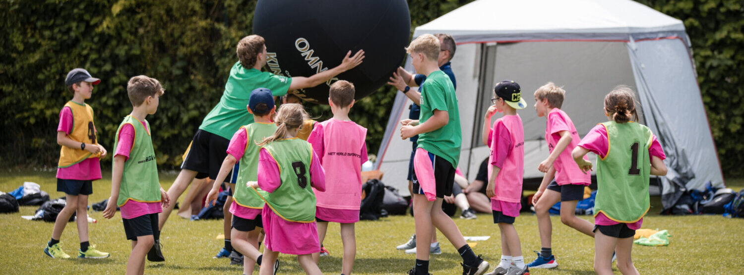 A group of children in pink and green t-shirts. They are playing Kinball. The giant black Kinball is above their heads which some of the children are reaching to touch