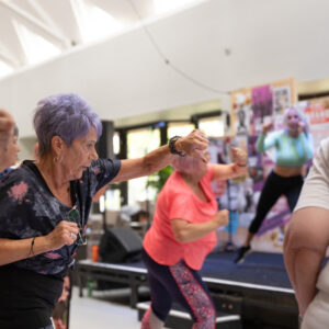 A group of older women people taking part in an exercise class. They're facing an instructor. They're doing high punches.