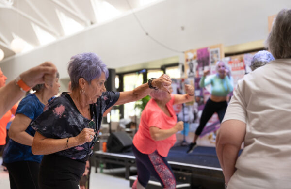 A group of older women people taking part in an exercise class. They're facing an instructor. They're doing high punches.