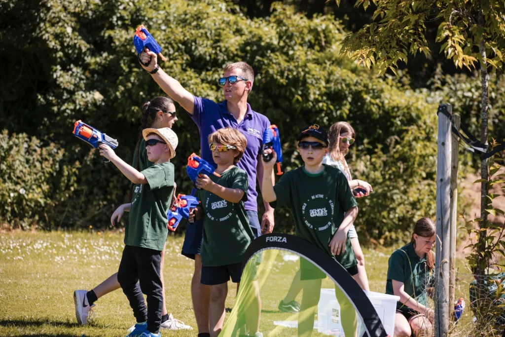 A group of young children engaging in active play. They are using nerf guns on a sunny day on a field.