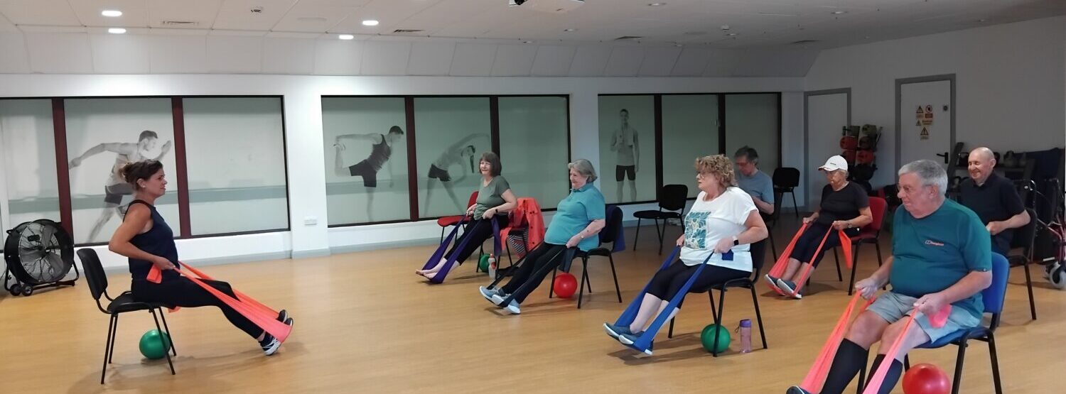A steady and strong class. The instructor is at the front of the room with rows of older adults on chairs in front. They are using a band to replicate a rowing movement.