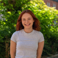 A woman with reddish hair wearing a light, grey top, smiling. Outside.