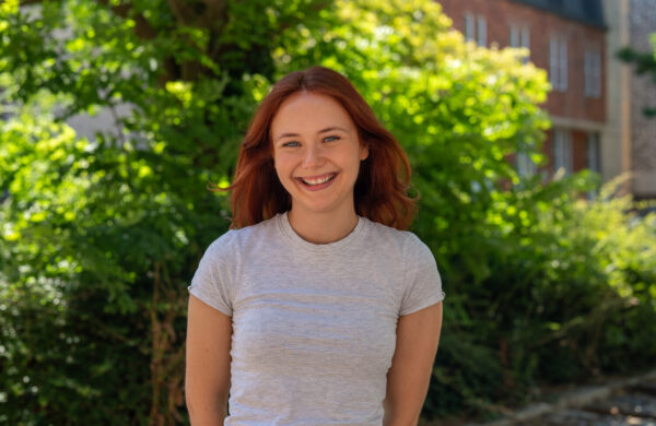 A woman with reddish hair wearing a light, grey top, smiling. Outside.
