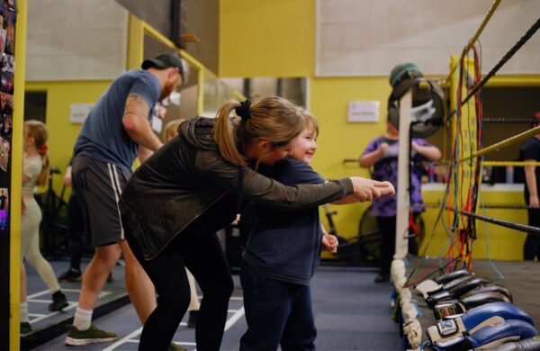 Hampshire Active Health participants. A mother and daughter in a boxing gym. The mother is leaning over the daughter, helping her to move her arm in a forward motion.