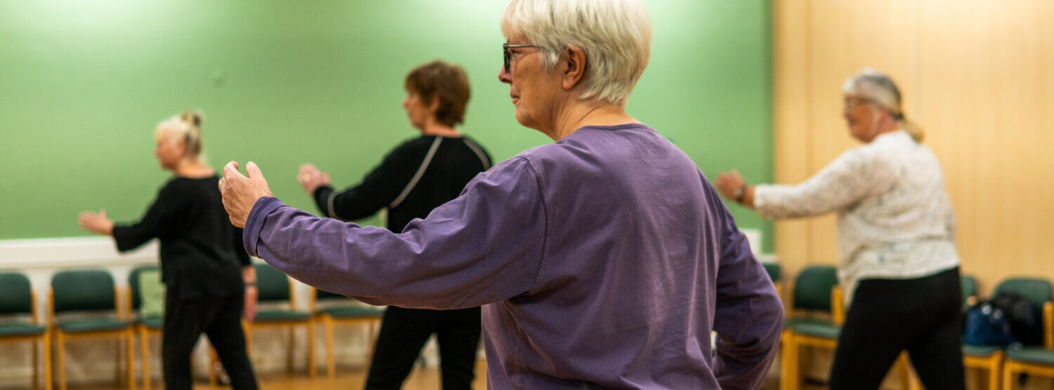A tai chi class. An older woman in a purple top is in the foreground.