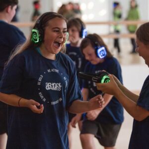 A group of young people in blue tshirts taking part in a silent disco. They are wearing headphones that are lit up. They have big smiles on their faces.