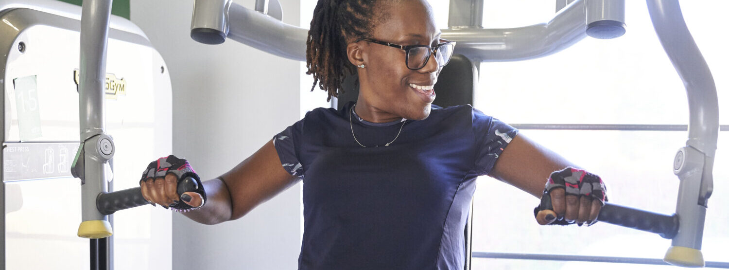 A woman on a gym machine that works out arms and chest. She is looking away from the camera smiling