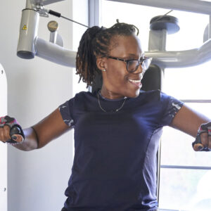 A woman on a gym machine that works out arms and chest. She is looking away from the camera smiling