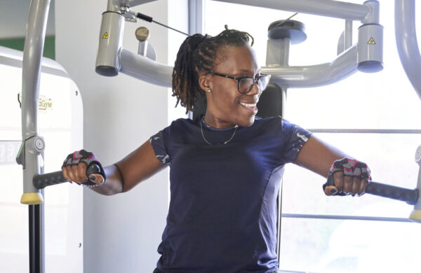 A woman on a gym machine that works out arms and chest. She is looking away from the camera smiling