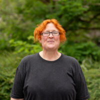 A woman with ginger hair and glasses, smiling. Outside.