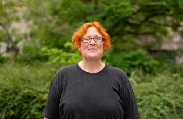 A woman with ginger hair and glasses, smiling. Outside.