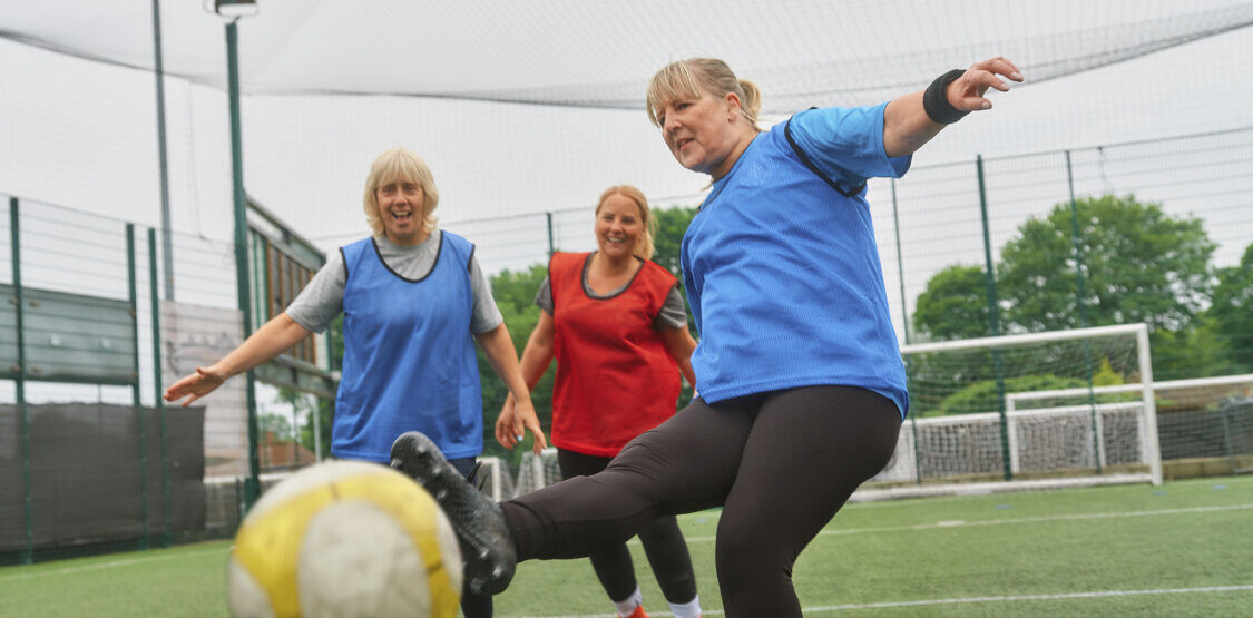 A group of older ladies playing football together on an artificial pitch. One is kicking the ball towards the camera smiling
