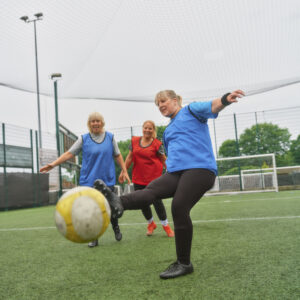 A group of older ladies playing football together on an artificial pitch. One is kicking the ball towards the camera smiling