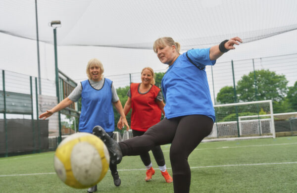 A group of older ladies playing football together on an artificial pitch. One is kicking the ball towards the camera smiling