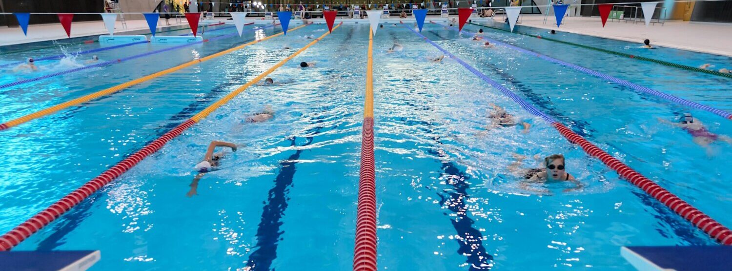 A swimming pool with people swimming in lanes. There is bunting across the top of the pool.