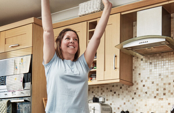 A women stands in her kitchen with her arms raised stretching tall. She is smiling