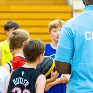 A group of young people facing a coach. One is holding a basketball and wearing a black and yellow basketball jersey. The others are dress in bright t-shirts - red, blue and yellow