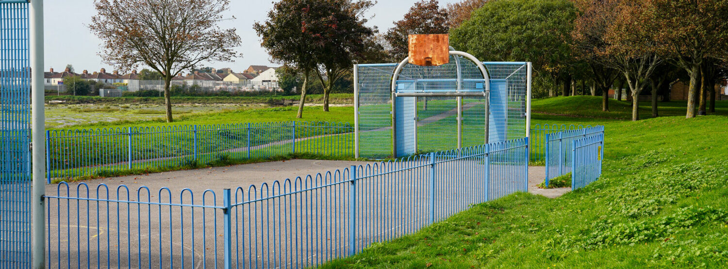 An outdoor concrete basketball court, surrounded by blue railings. Its surrounded by grass and overlooked by homes.