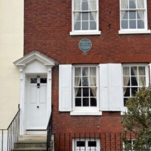 A red brick terraced building with a white door. A small plaque beneath the window.