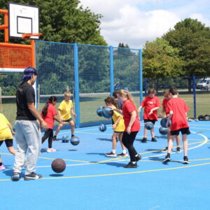An outdoor basketball court. Children in red and yellow t-shirts practice moving while bouncing the ball.