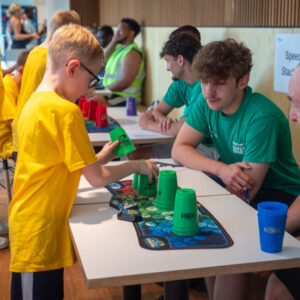 A group of children stood in front of a table with green cups on it, speed stacking.