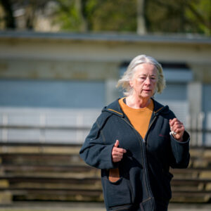 A woman with grey hair in a black zip up sweatshirt running outdoors.